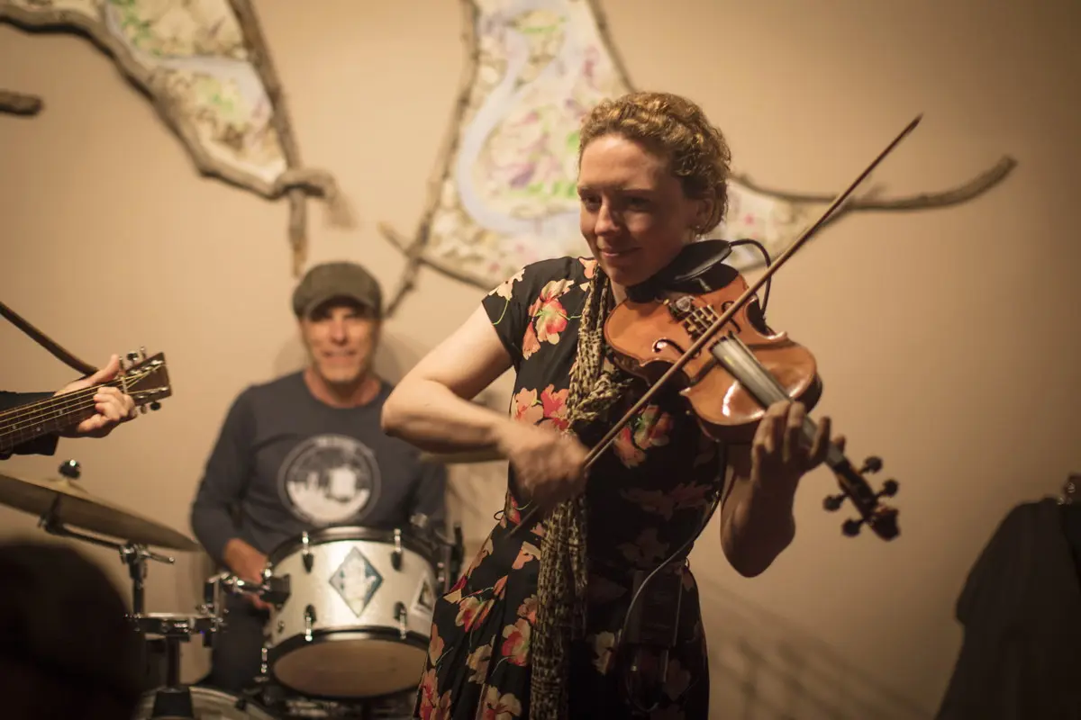 A musician playing violin with a guitarist and drummer in the background.