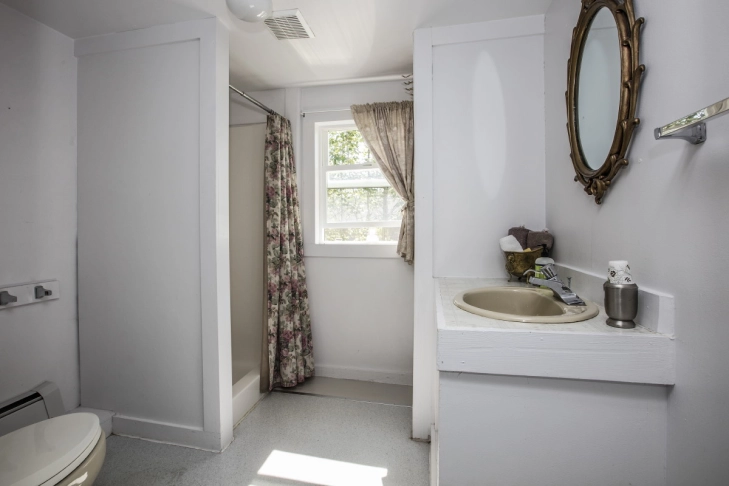 Small bathroom with a toilet, sink, window with curtains, and an oval mirror.