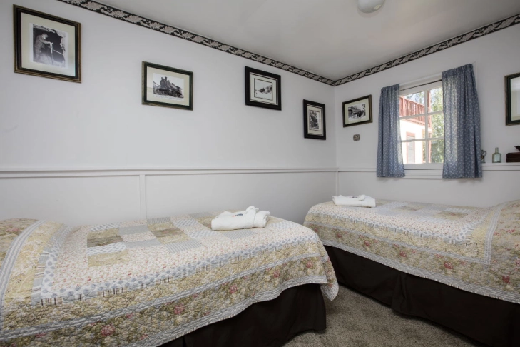A cozy twin bedroom with patterned quilts, framed pictures on the wall, and a window with blue curtains.