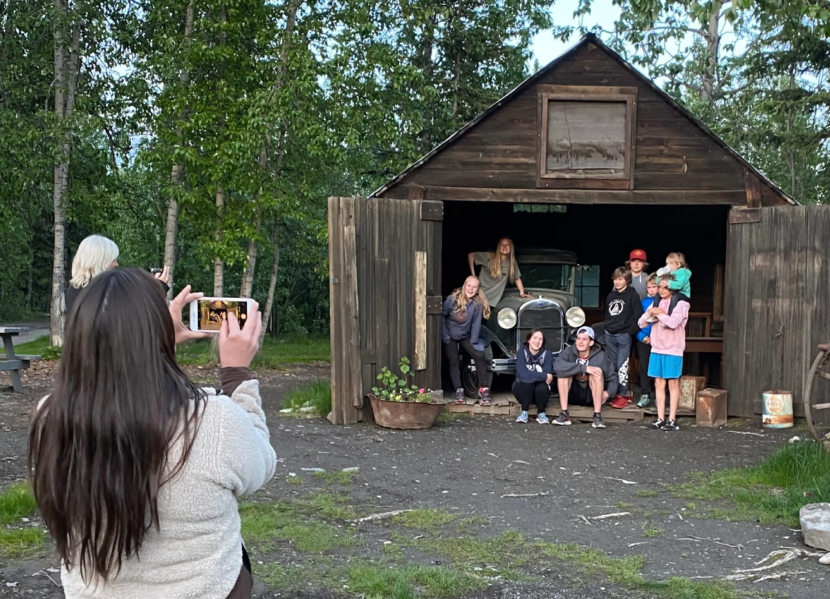A person taking a photo of a group in a rustic wooden garage with an old car